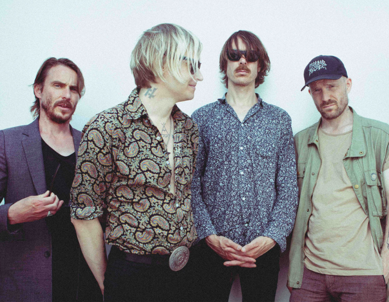 Imagem de quatro integrantes da banda Refused posando lado a lado. Eles estão vestidos com estilos variados que combinam elementos casuais e alternativos, como camisas estampadas, jaquetas e acessórios. O fundo é neutro, destacando o visual dos músicos.