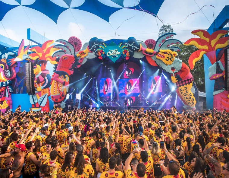 Multidão de foliões vestindo camisetas amarelas em festa ao ar livre, diante de um palco decorado com elementos coloridos e tema tropical com inscrição 'Carnaval na cidade', luzes de show e céu azul ao fundo.